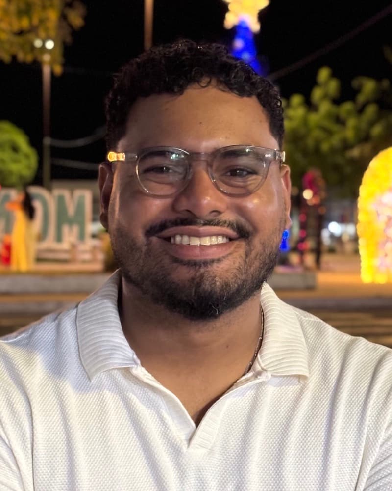 Jorge Eder sorrindo, de óculos e camisa polo branca, numa praça iluminada à noite